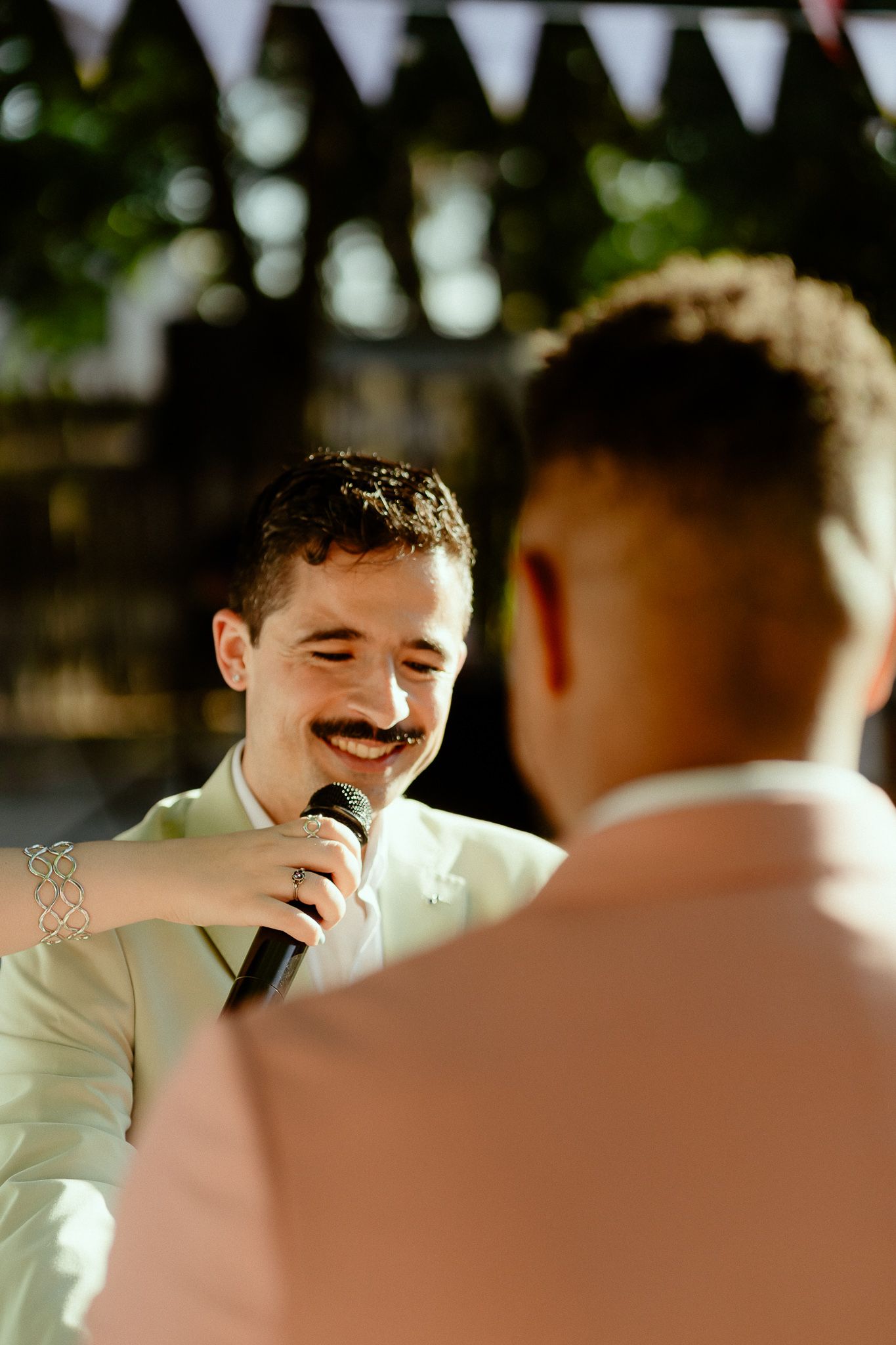 Man with mustache smiling while holding microphone at outdoor wedding reception with bunting decorations.