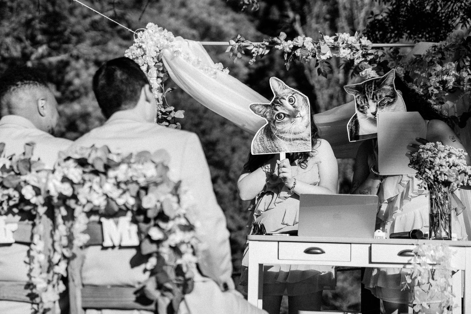Black and white outdoor wedding ceremony with guests wearing animal masks seated at a decorated table with floral arrangements.