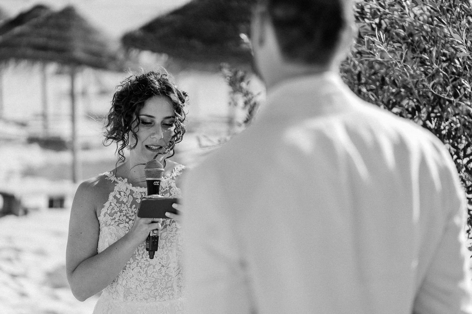 Bride reads wedding vows from a card during an outdoor beach ceremony in a lace dress, black and white photo.