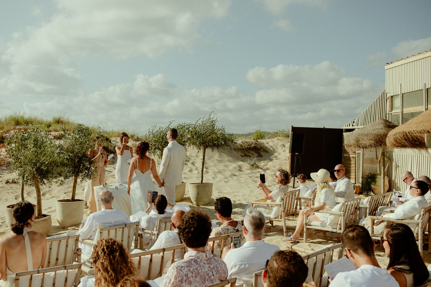 Outdoor beach wedding ceremony with guests seated on wooden chairs, couple at altar near sand dunes on a sunny day.