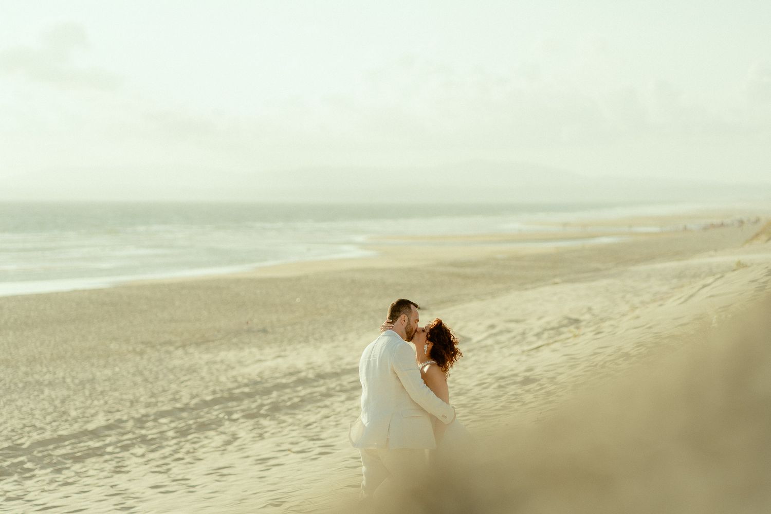 Couple sharing a romantic kiss on a serene sandy beach with ocean waves in the background.