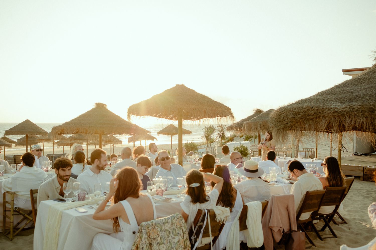 Outdoor beachside wedding reception at sunset with guests in white attire seated at elegant tables under thatched umbrellas.