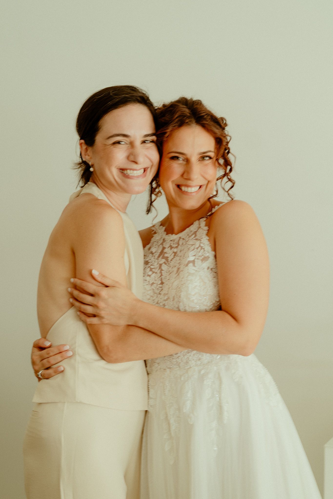 Bride in lace wedding dress hugging smiling woman in cream outfit against light background.