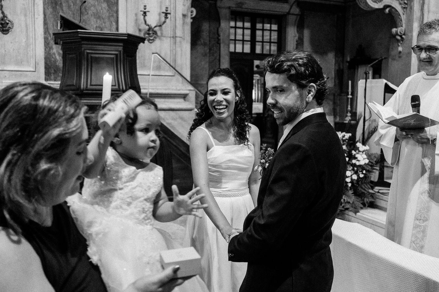 Black and white wedding ceremony photo with bride, groom, and flower girl sharing a joyful moment inside a church.