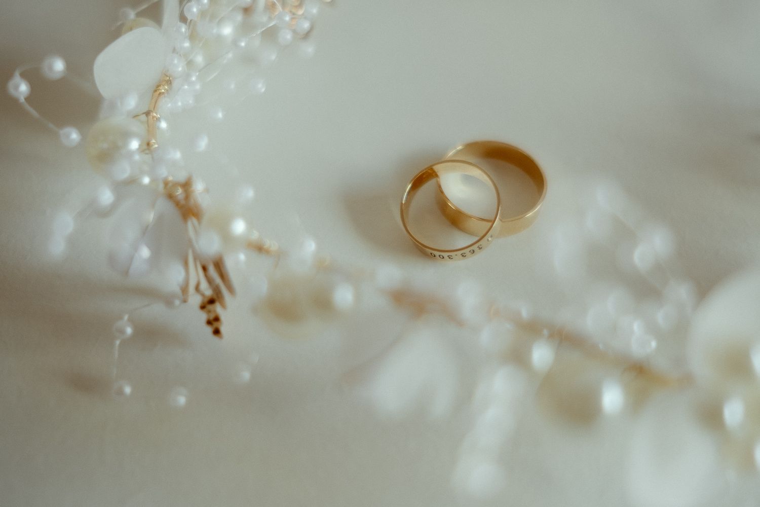 Two gold wedding rings resting on a white floral arrangement with delicate pearls and dried flowers.