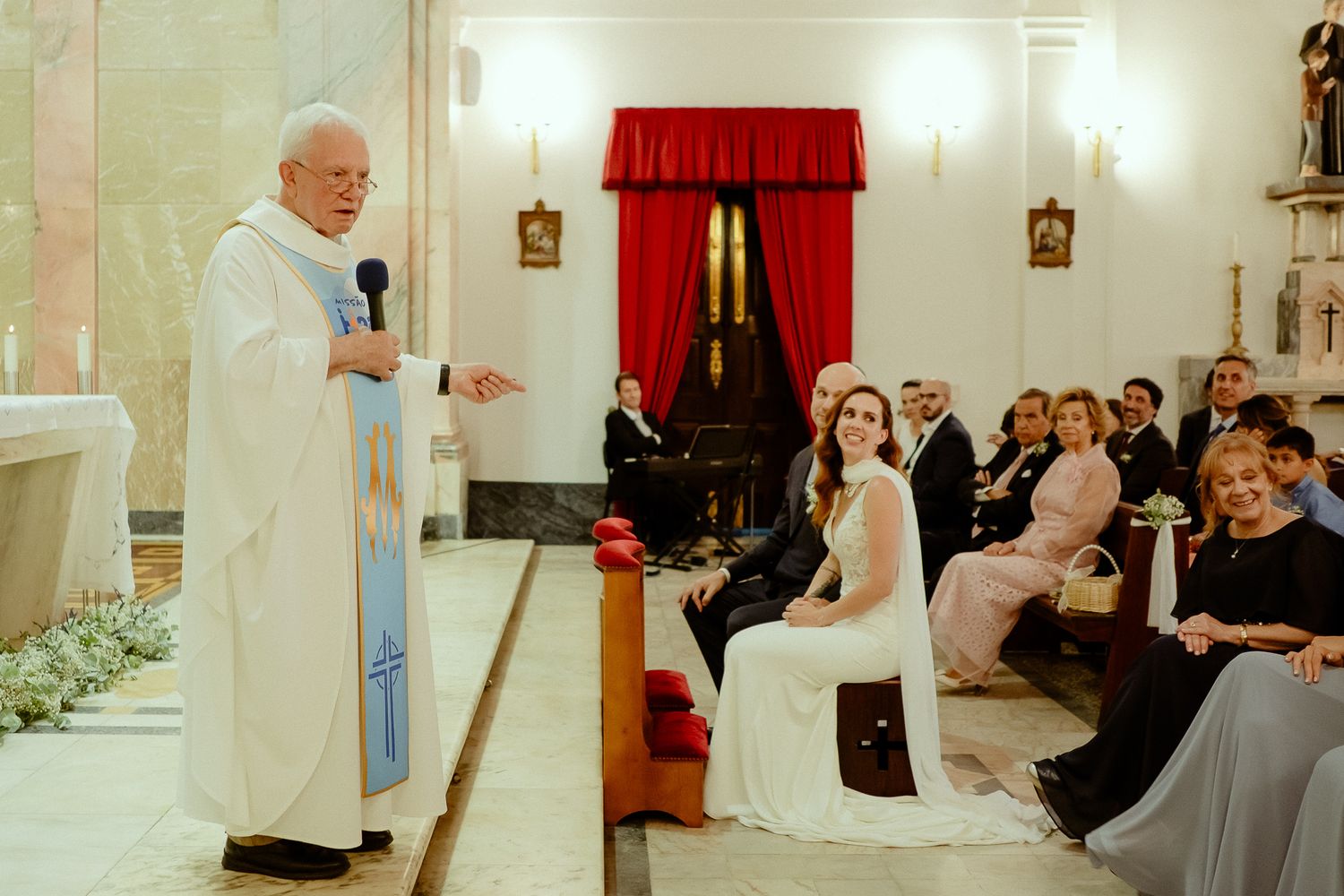 Priest addresses bride and wedding guests during traditional church ceremony with white floral decor.