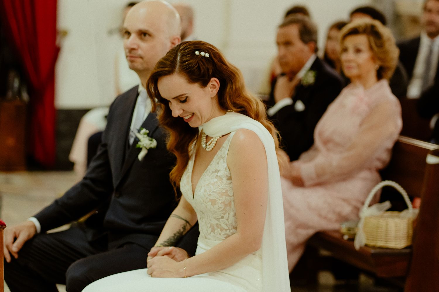 Bride in white wedding gown smiling during church ceremony, seated on pew with guests in background.