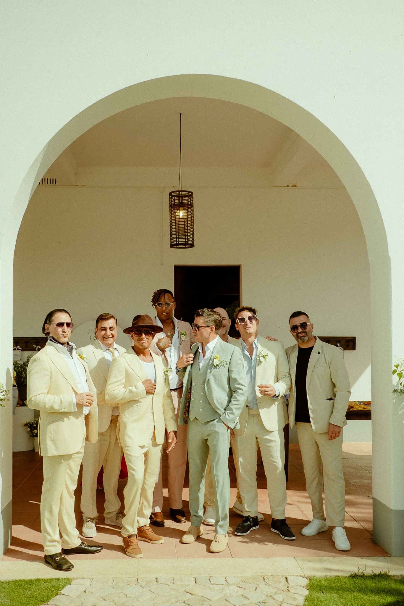 Groom and groomsmen in cream suits pose under a white arch at an outdoor wedding venue.