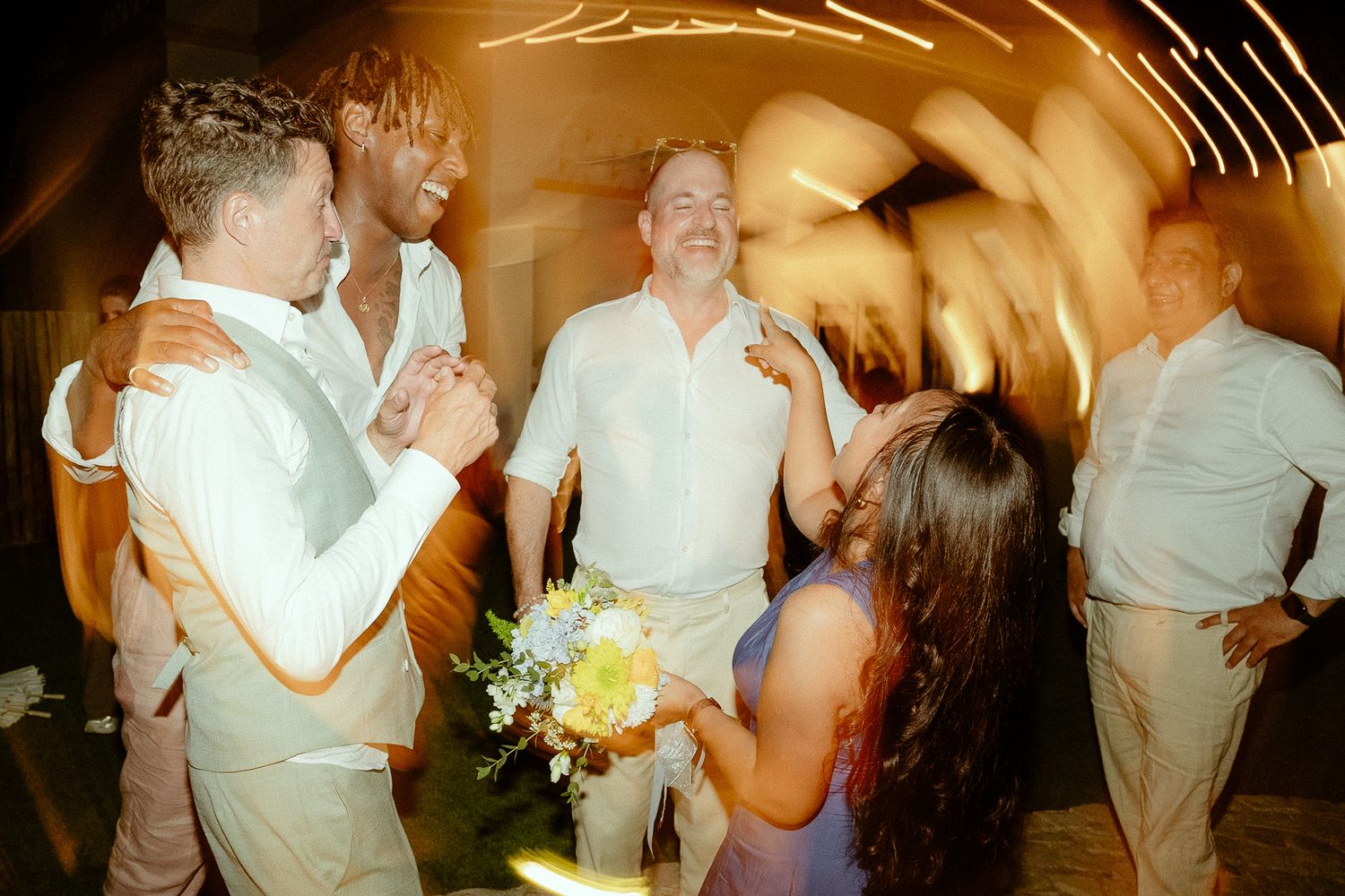 Wedding guests celebrate joyfully, with a bride holding a bouquet surrounded by smiling friends at a warm, festive reception.