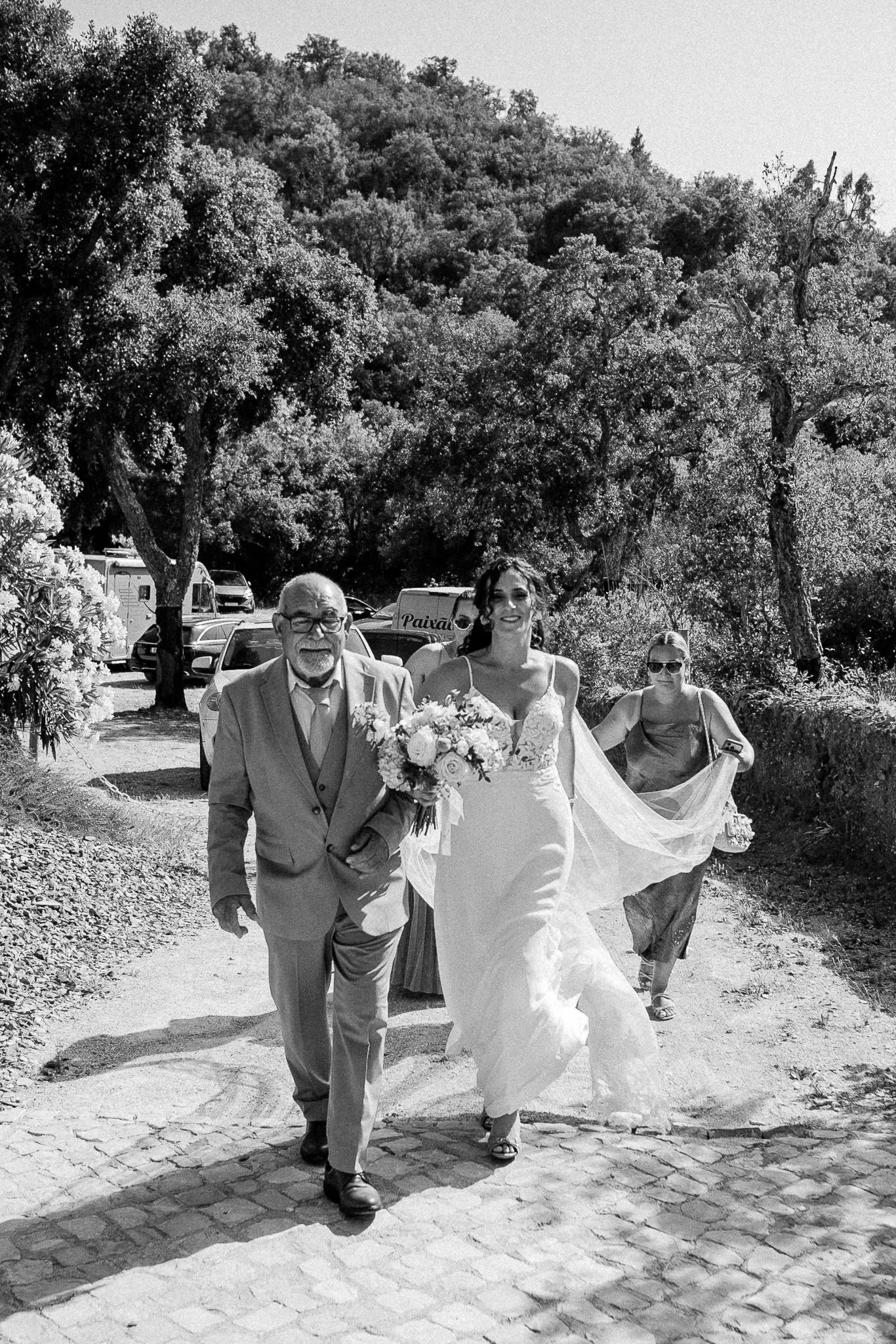 Black and white photo of a bride walking with her father and bridesmaid on a cobblestone path at an outdoor wedding.