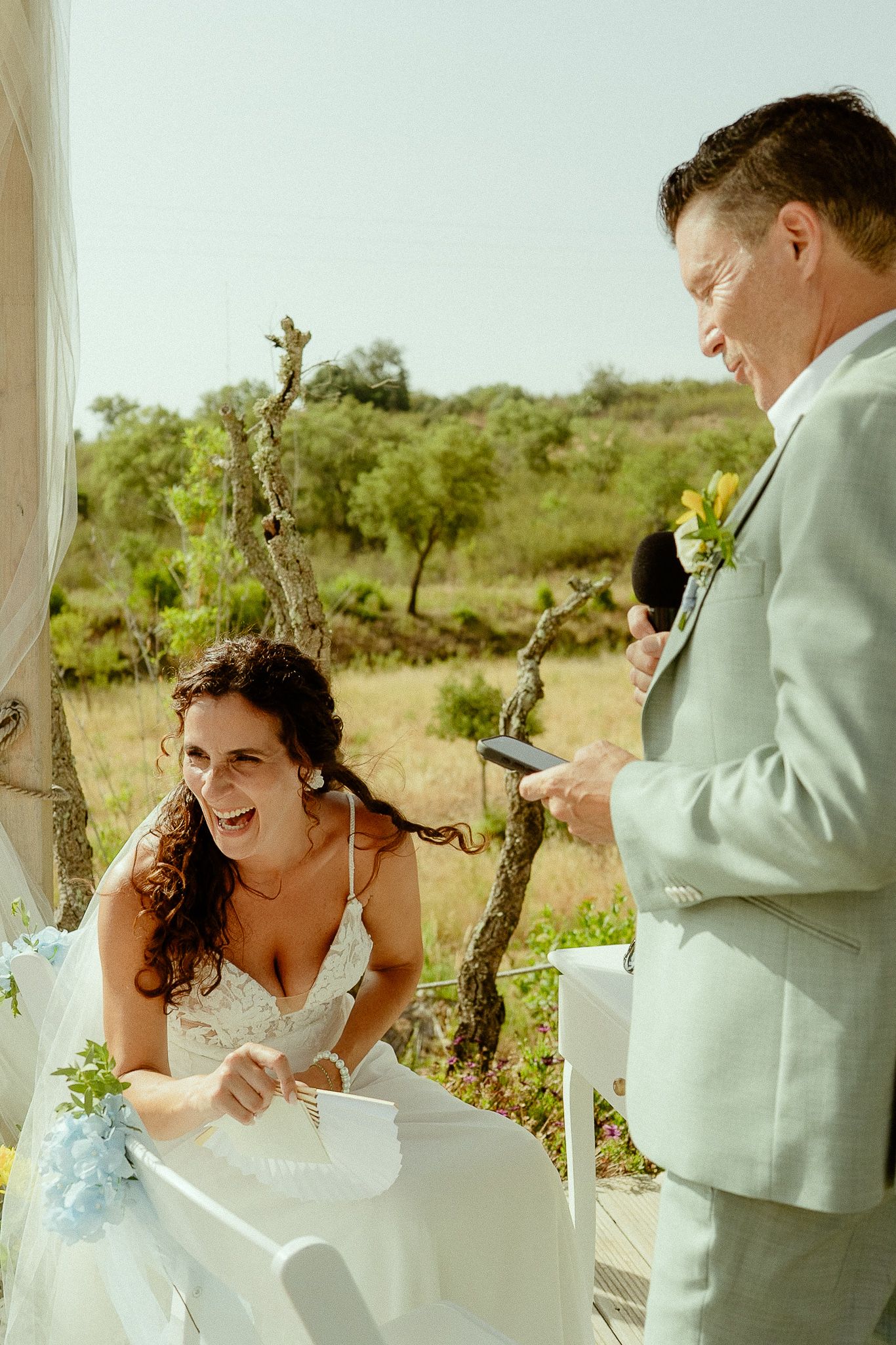 Bride laughing joyfully with groom in light suit during outdoor wedding ceremony surrounded by natural greenery.