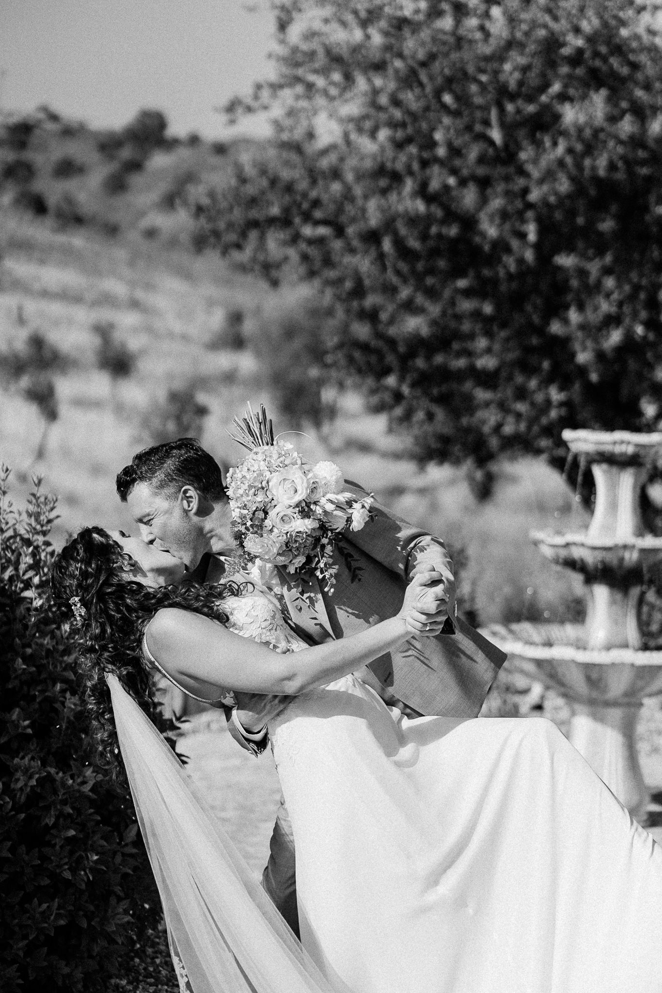 Black and white photo of a groom dipping and kissing his bride holding a bouquet near a fountain outdoors.