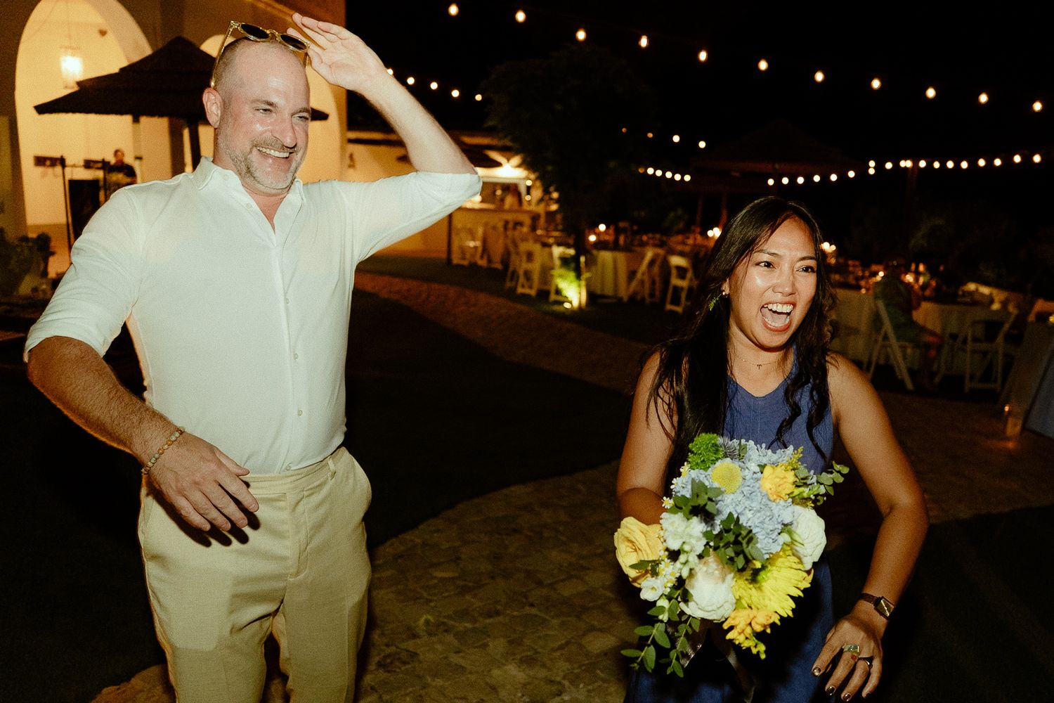 A man and woman laughing outdoors at a night event, woman holding yellow and white bouquet under string lights.