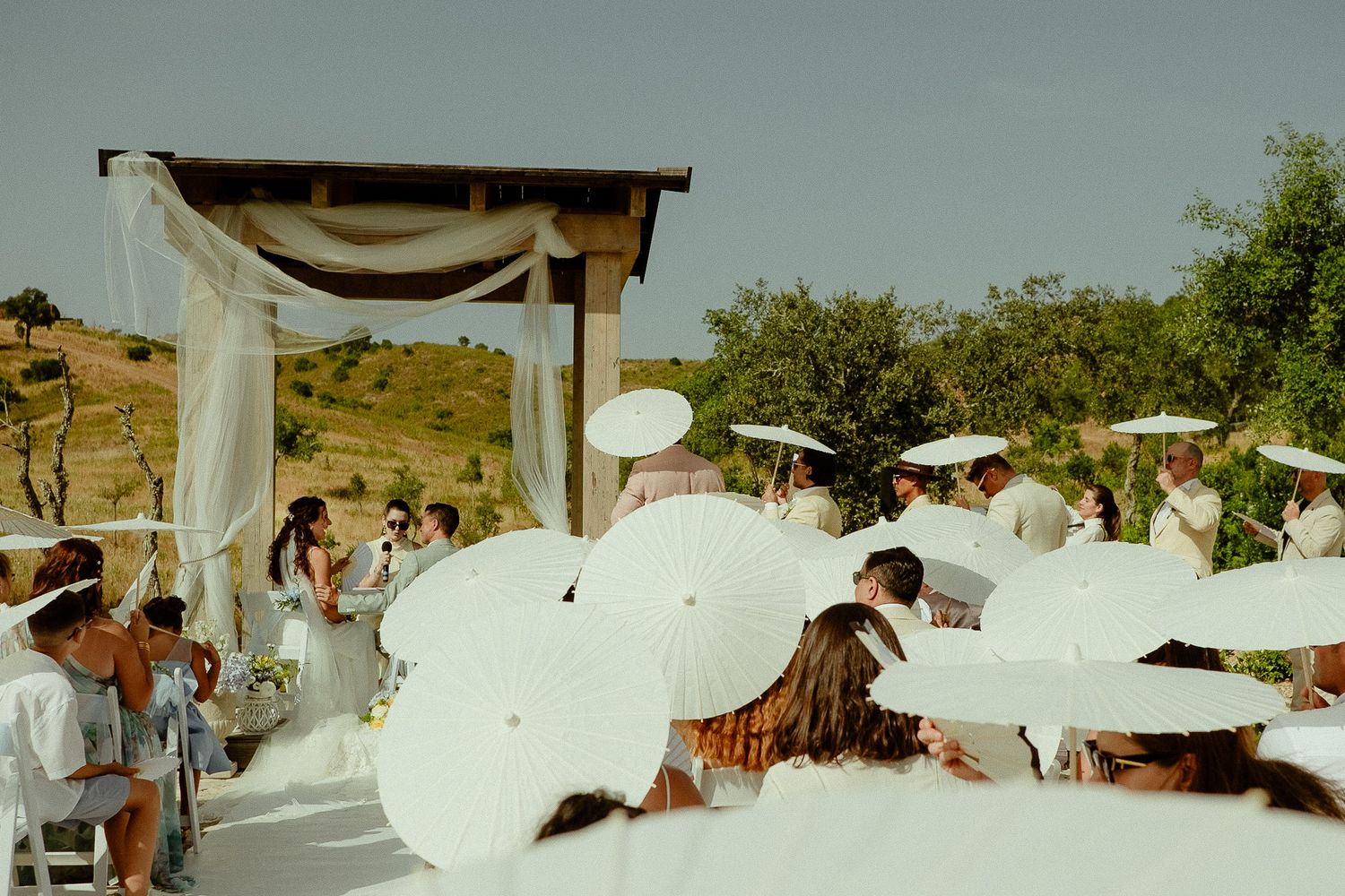 Outdoor wedding ceremony with guests holding white parasols under a sunny sky near a draped altar.