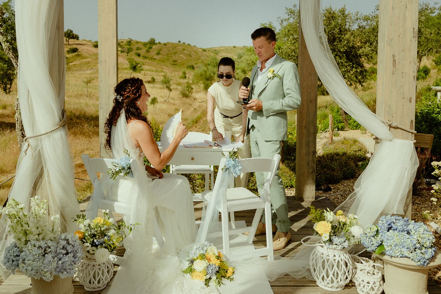 Outdoor wedding ceremony with bride, groom, and officiant under a rustic arch with blue hydrangeas and white draping.
