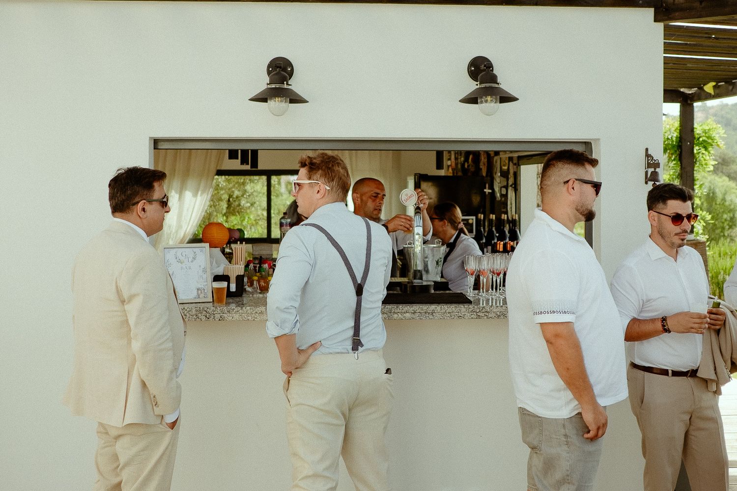 Men in smart casual attire gather at an outdoor bar during a wedding reception on a sunny day.