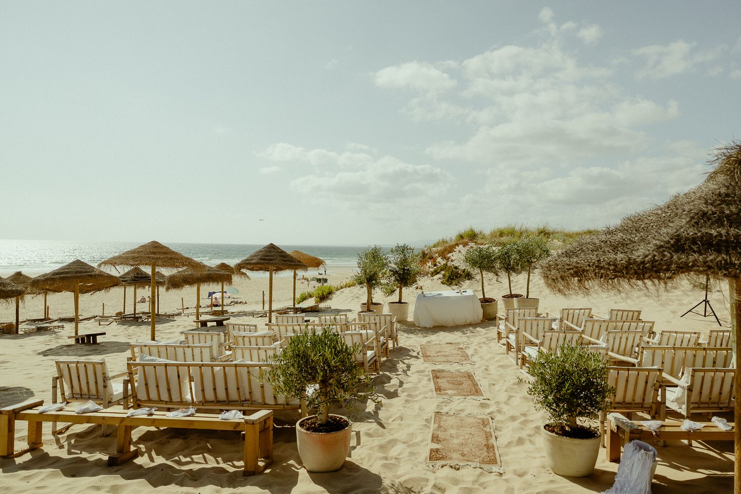 Beachside lounge area with straw umbrellas, white wooden chairs, and potted plants on sandy shore under sunny sky.