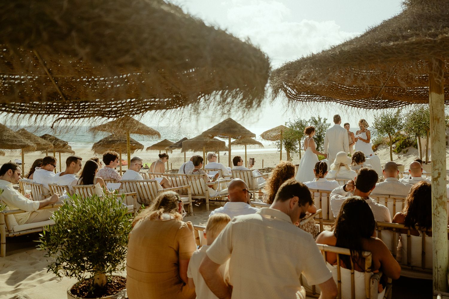 Guests gather at a beachside bar under thatched umbrellas with ocean views on a sunny day.