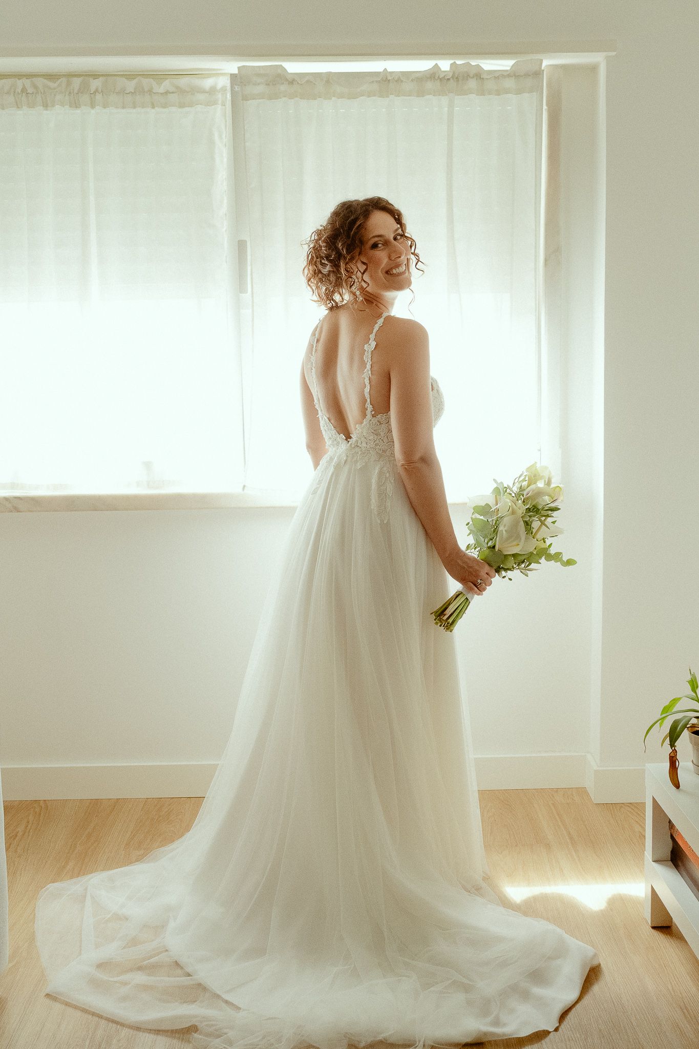 Bride in white lace tulle gown holding bouquet, smiling over shoulder near bright window.
