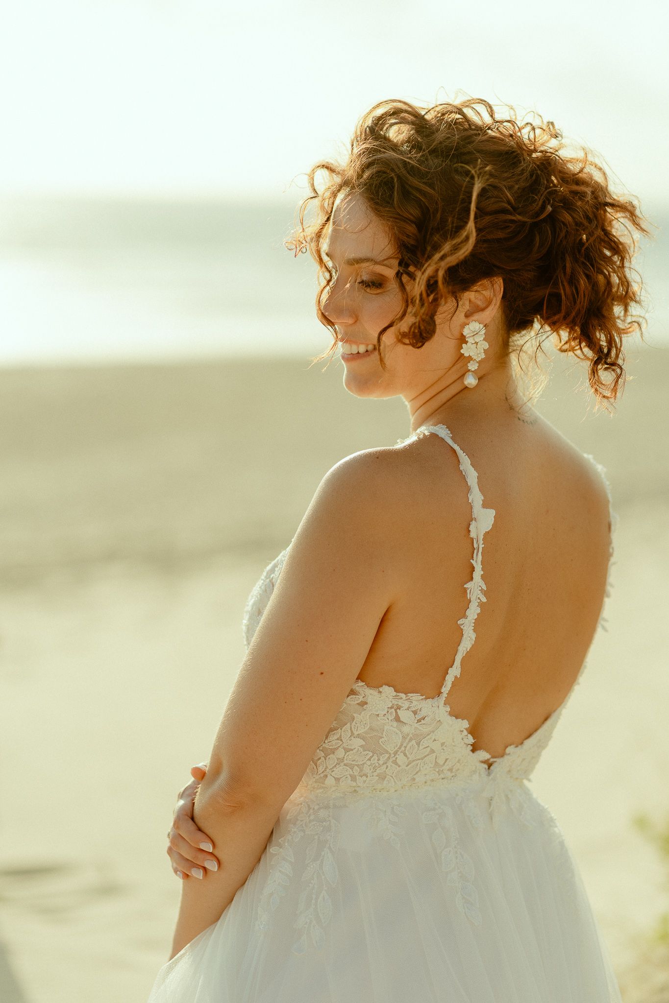 Smiling bride in backless white lace gown with curly updo and floral earrings on a sunny beach.