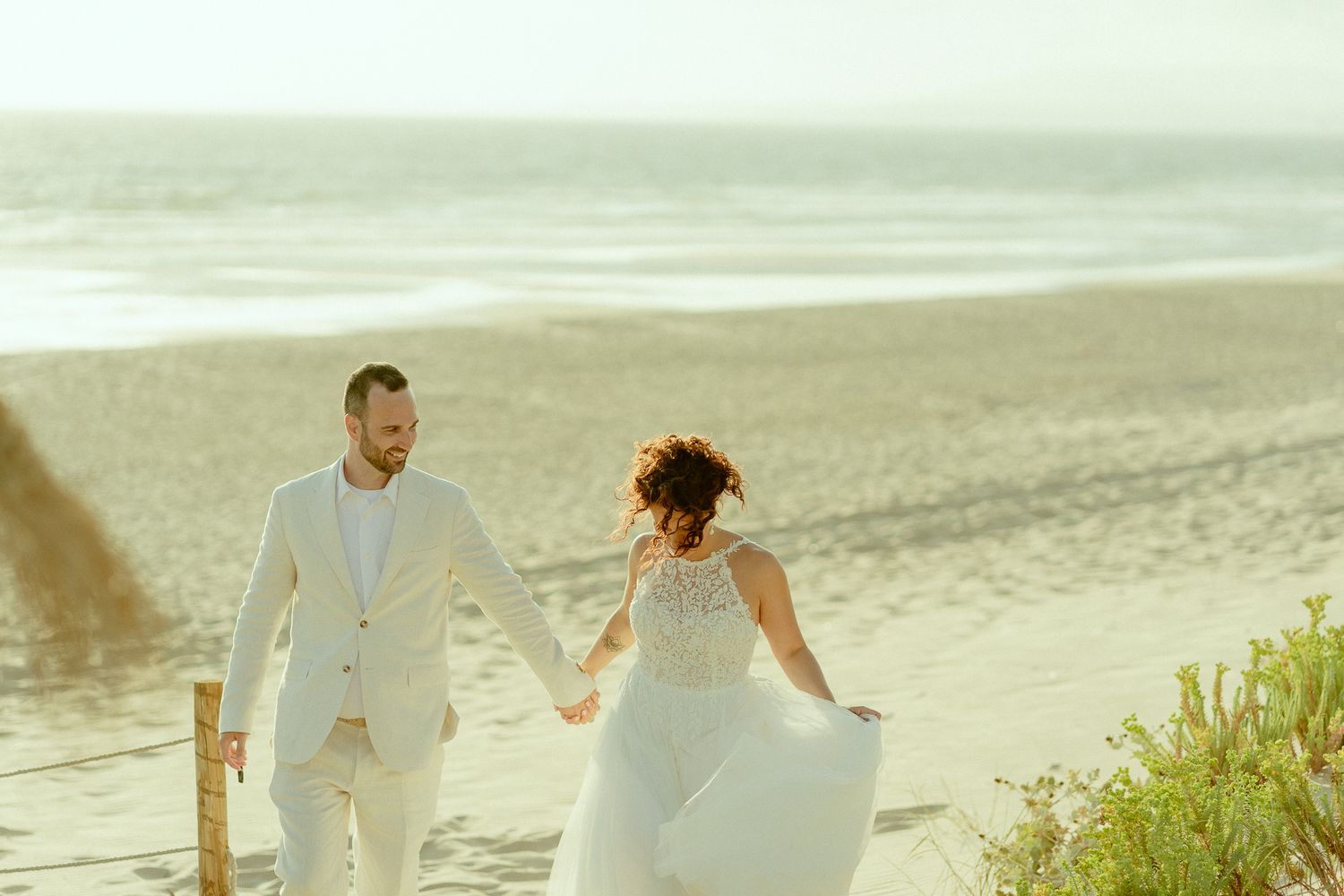 Bride and groom holding hands walking on a sandy beach in elegant white wedding attire with ocean waves in the background.