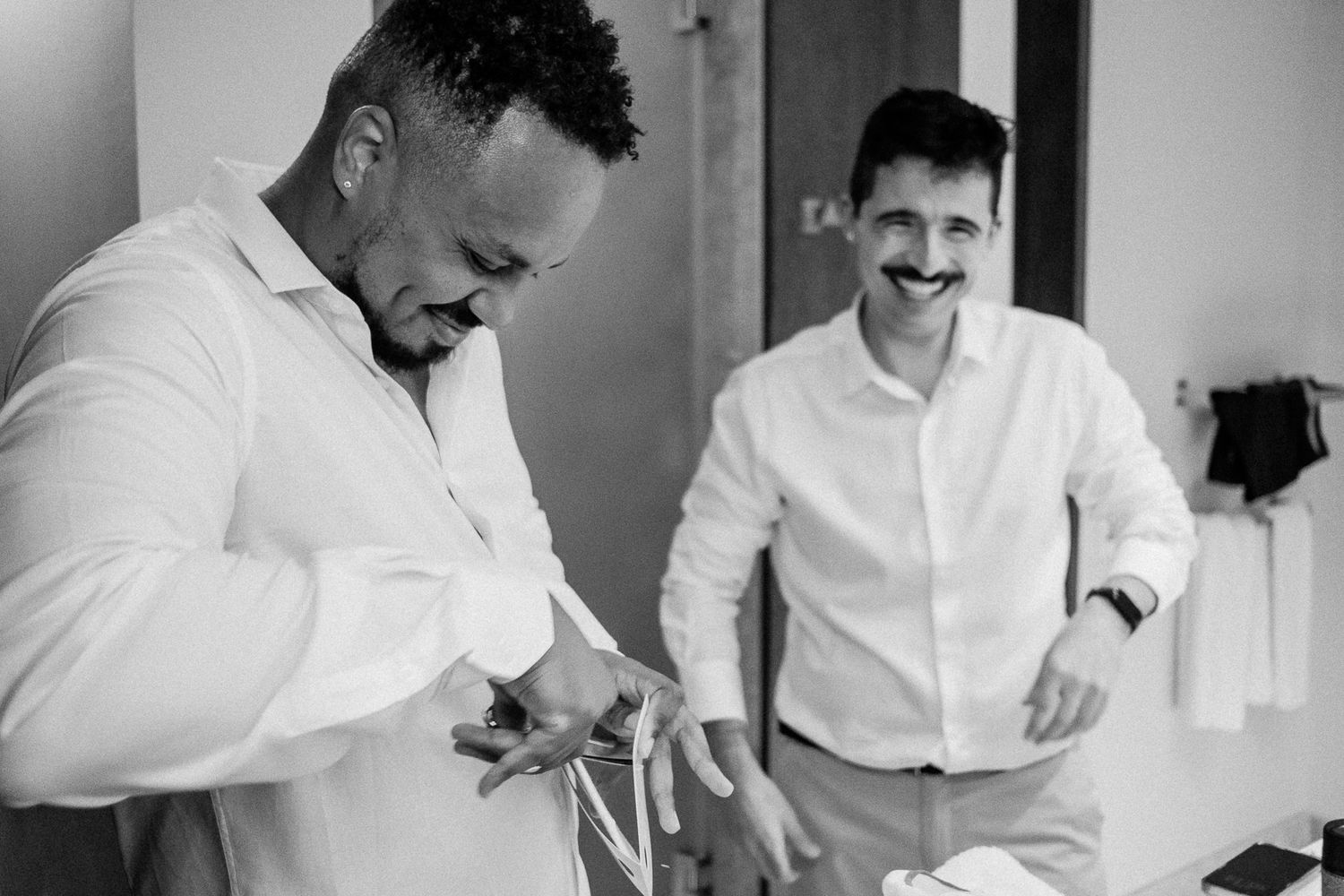 Two men in white shirts laugh while preparing for a wedding, one adjusting a tie in a black and white photo.