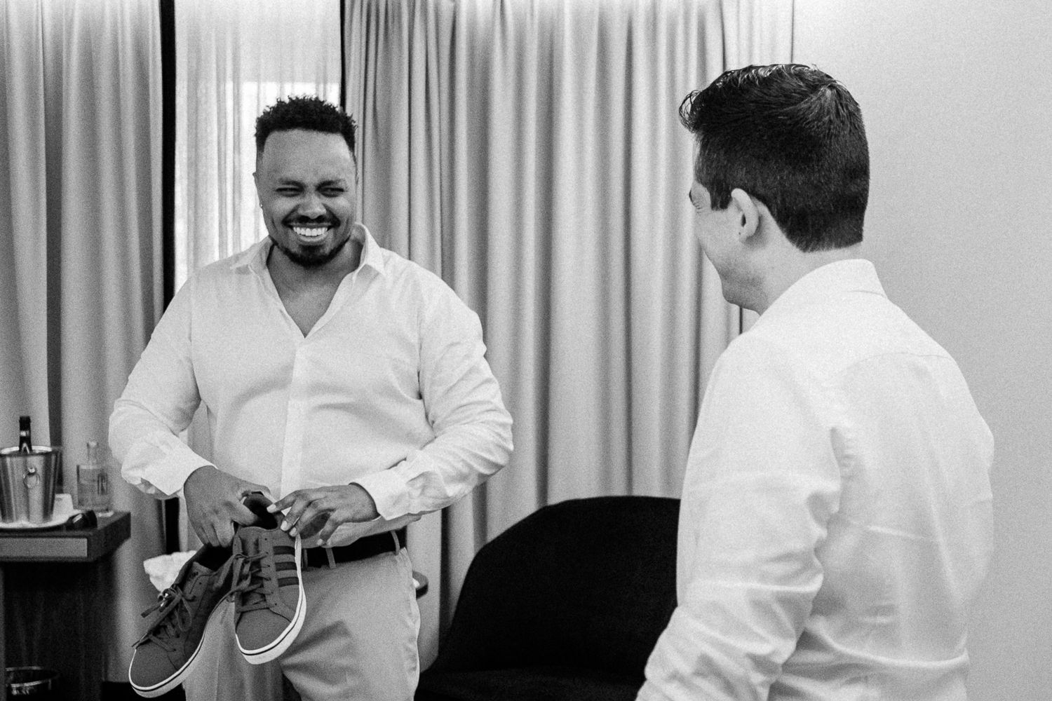 Two men in white shirts share a laugh, one holding shoes, in a black and white wedding preparation photo.
