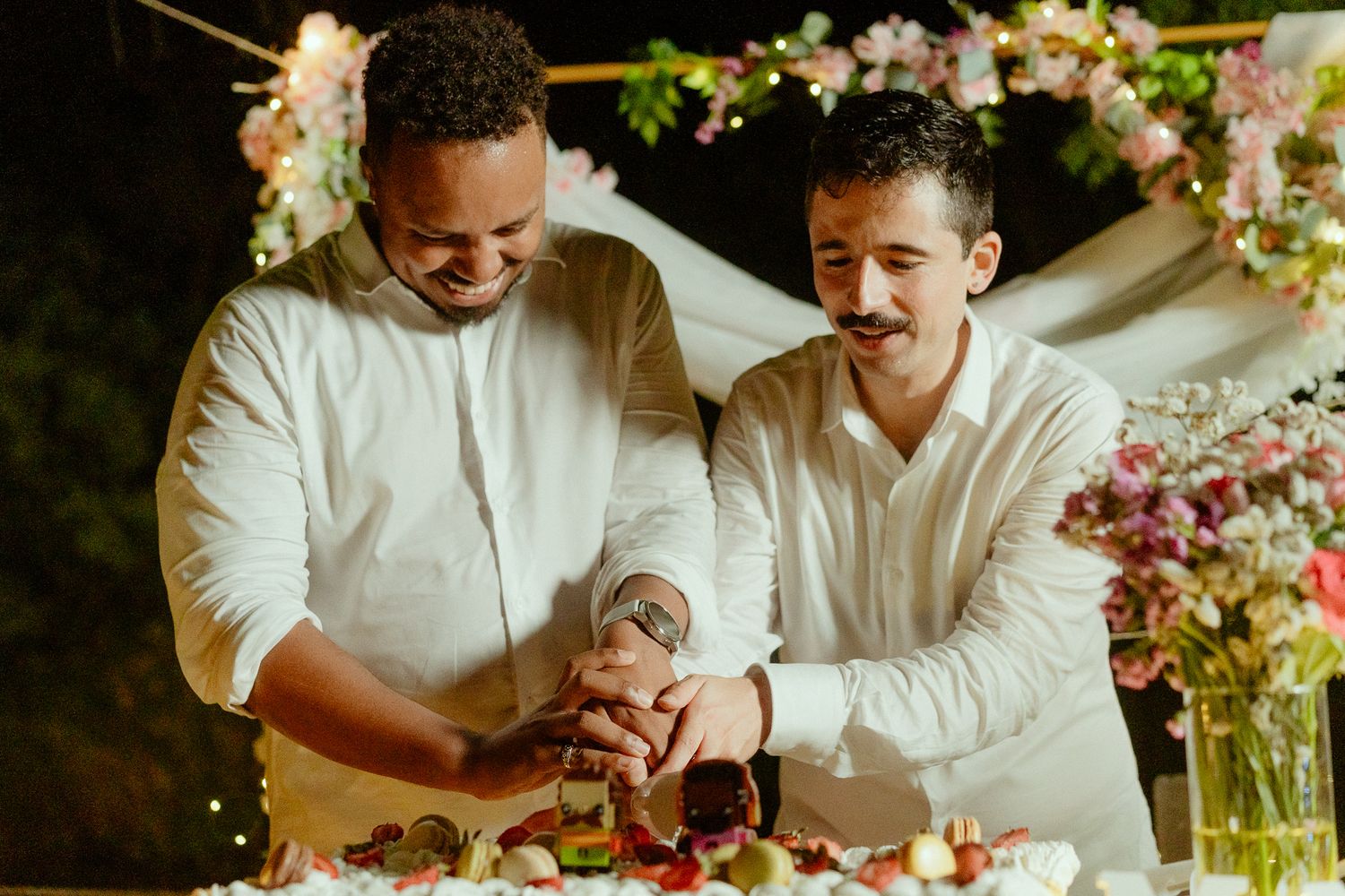 Two grooms cutting wedding cake together at floral-decorated outdoor reception at night.