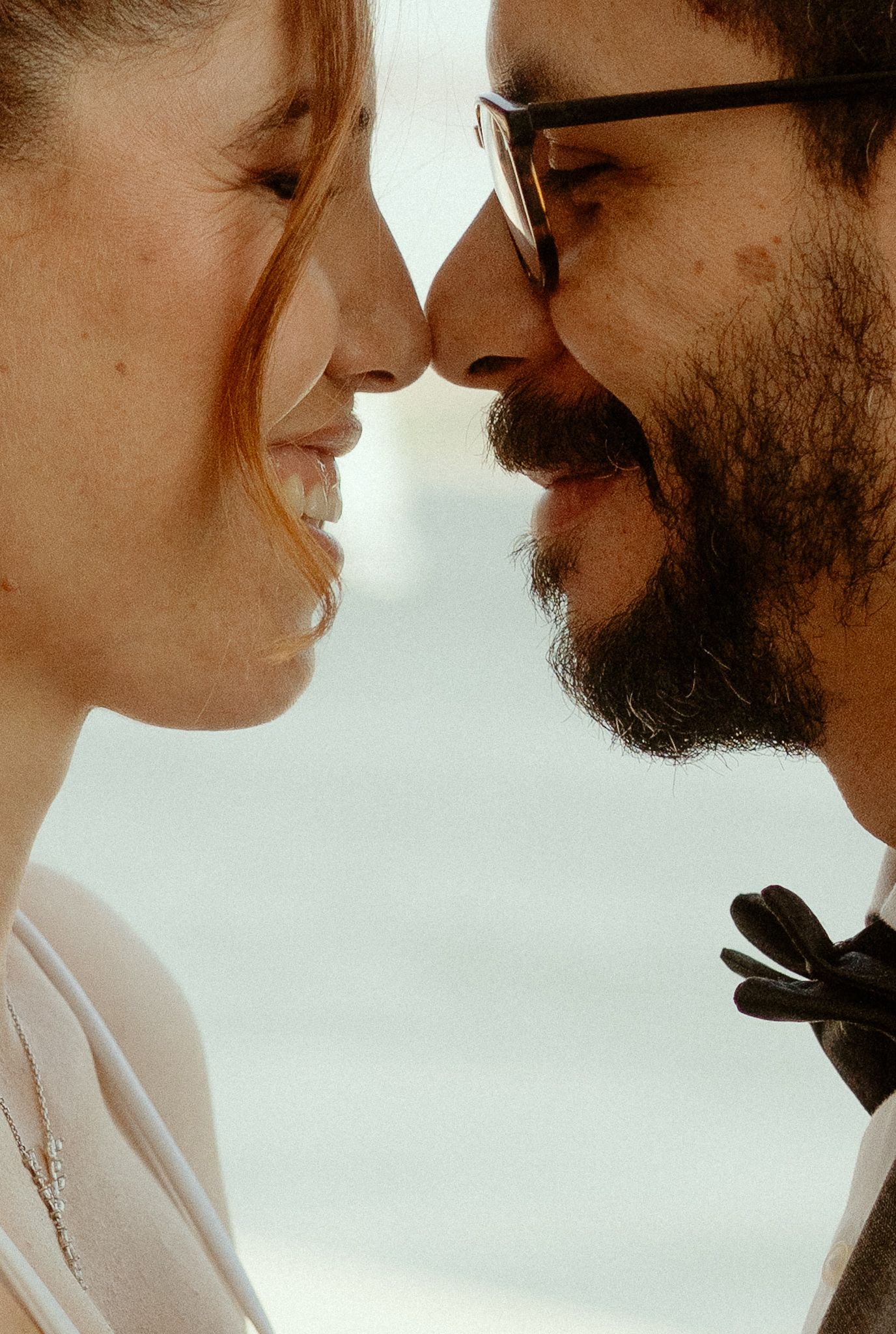 A couple with noses almost touching, sharing an intimate moment, the man wearing glasses and a beard.