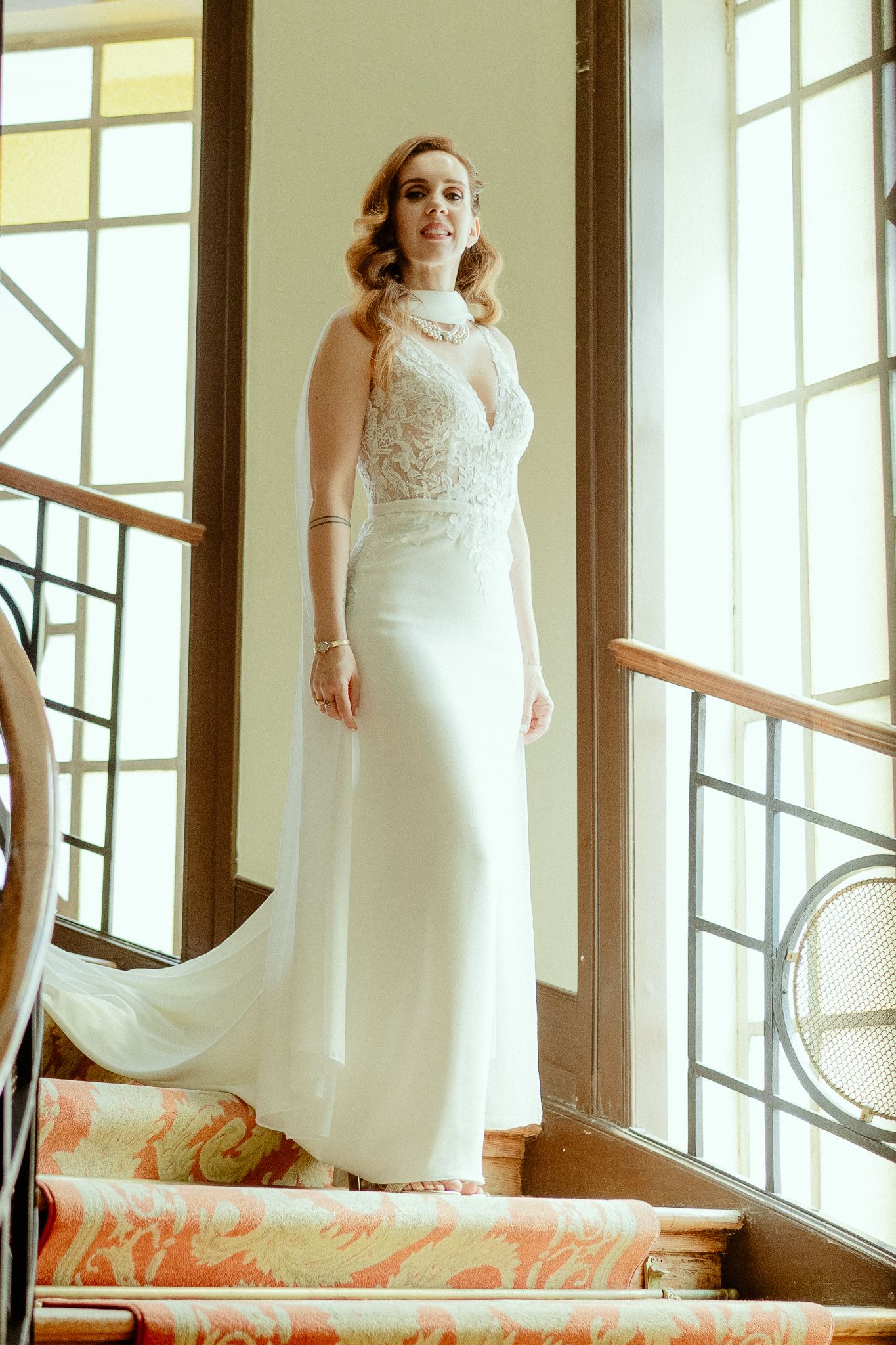 Bride in elegant white lace halter wedding gown standing on staircase by large windows in natural light.