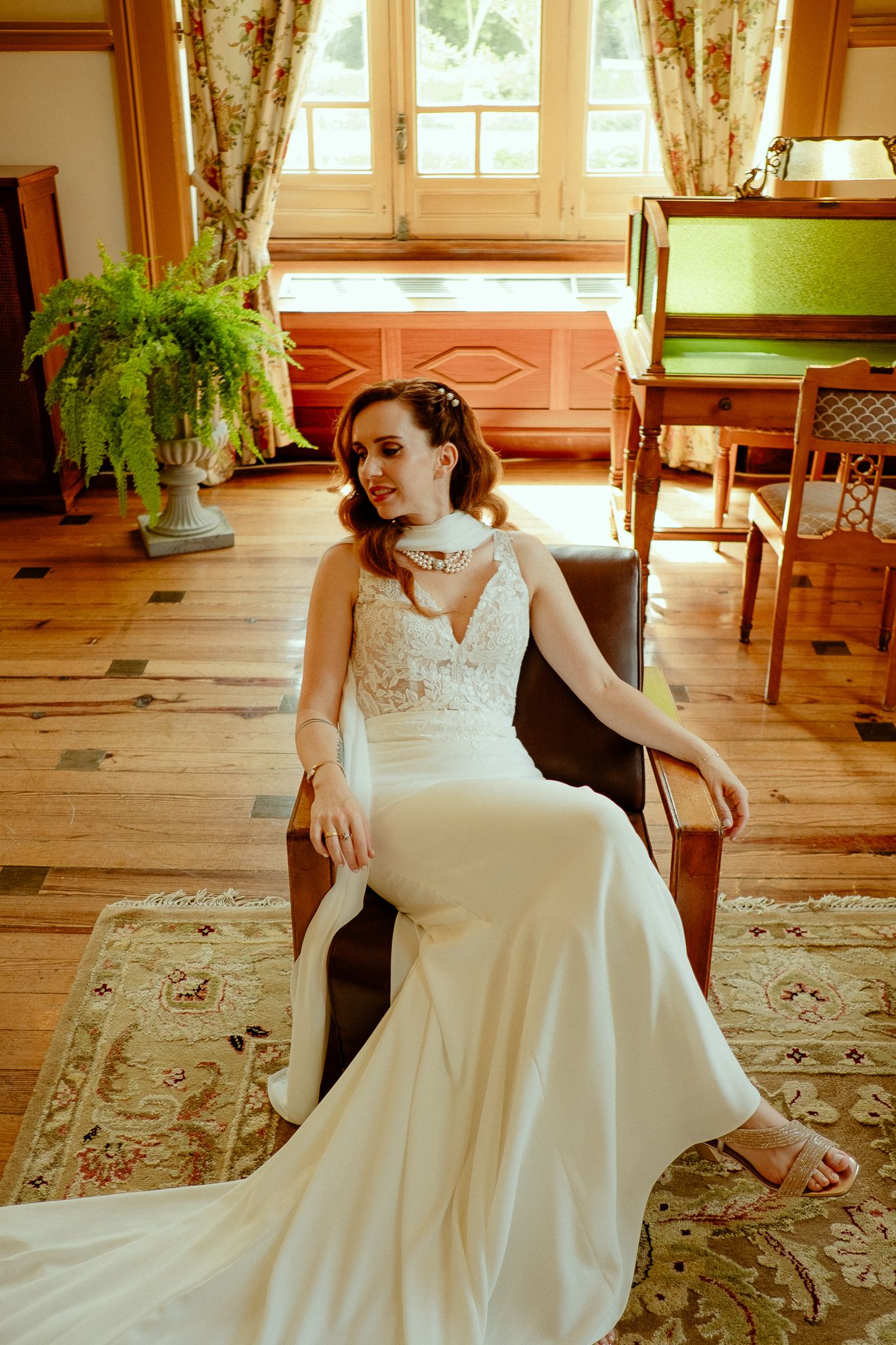 Bride in white lace gown seated in leather chair on patterned rug in elegant wood-floored room with fern and windows.