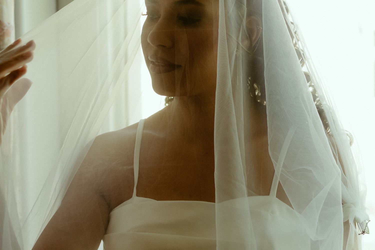 A bride in a white wedding dress and veil smiles softly in natural light during bridal preparations.