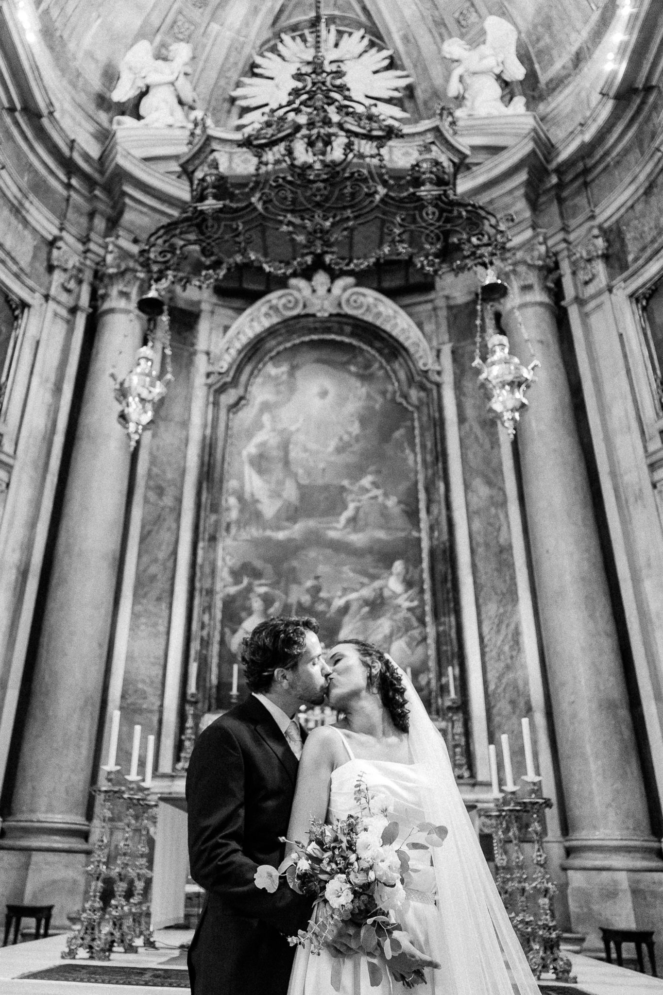 Bride and groom share a kiss inside an ornate baroque church with angel sculptures and a classical painting.