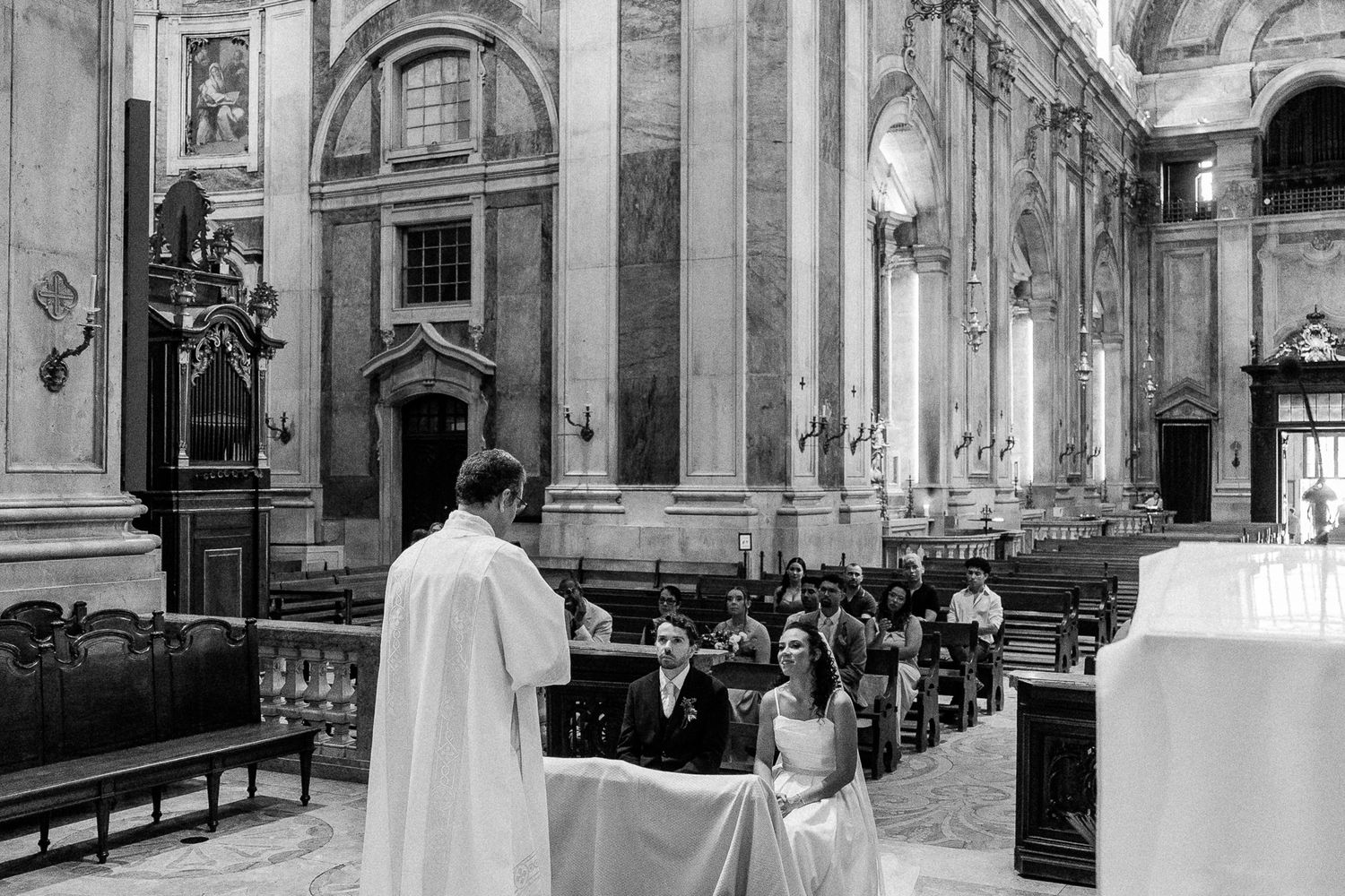 Black and white photo of a wedding ceremony inside an ornate church with guests seated in pews.