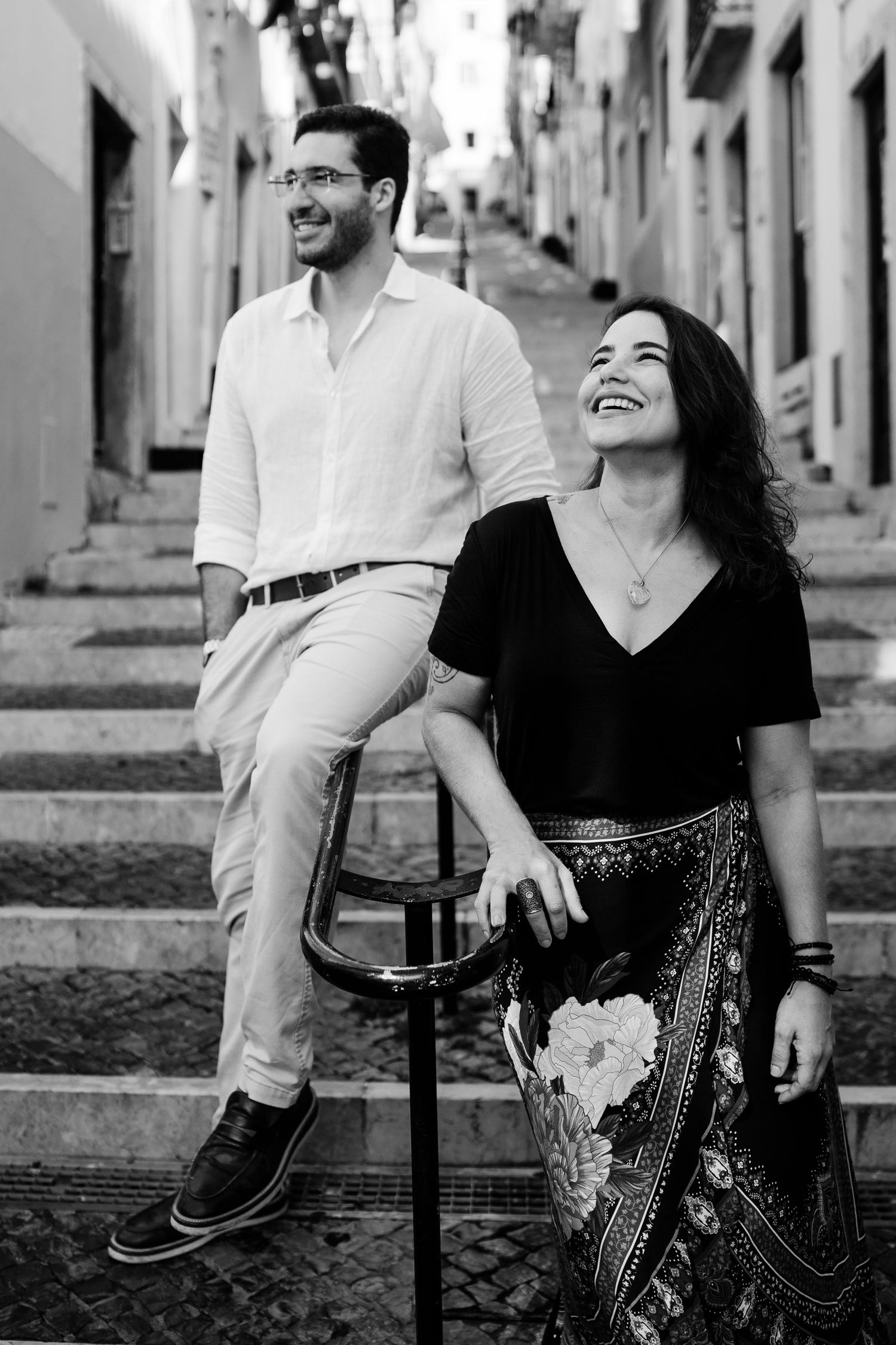 A happy couple laugh together while sitting on outdoor stairs in a charming black and white photo series.