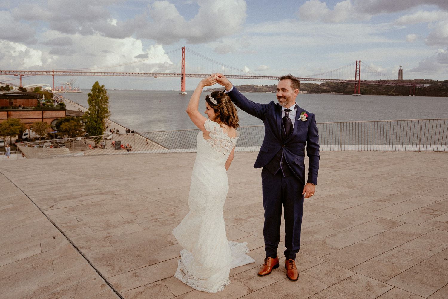 A wedding couple dances together on a scenic overlook with the 25 de Abril Bridge and Tagus River in Lisbon behind them.
