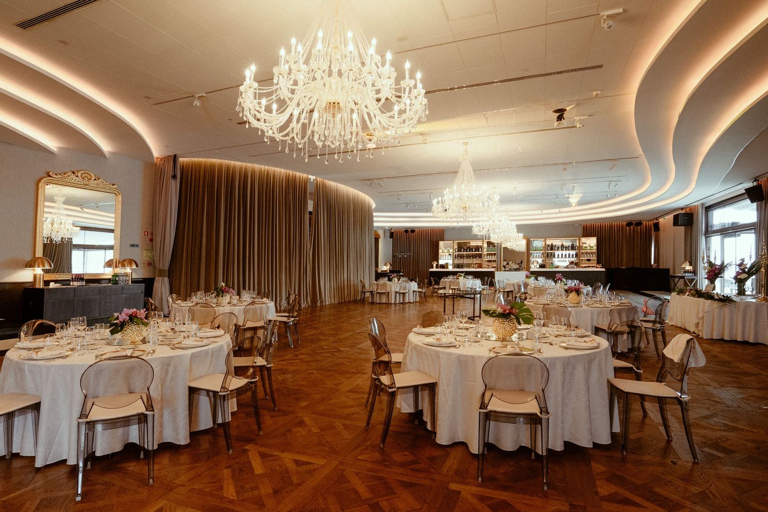 Elegant banquet hall with crystal chandelier, round tables with white linens, and herringbone hardwood floors.