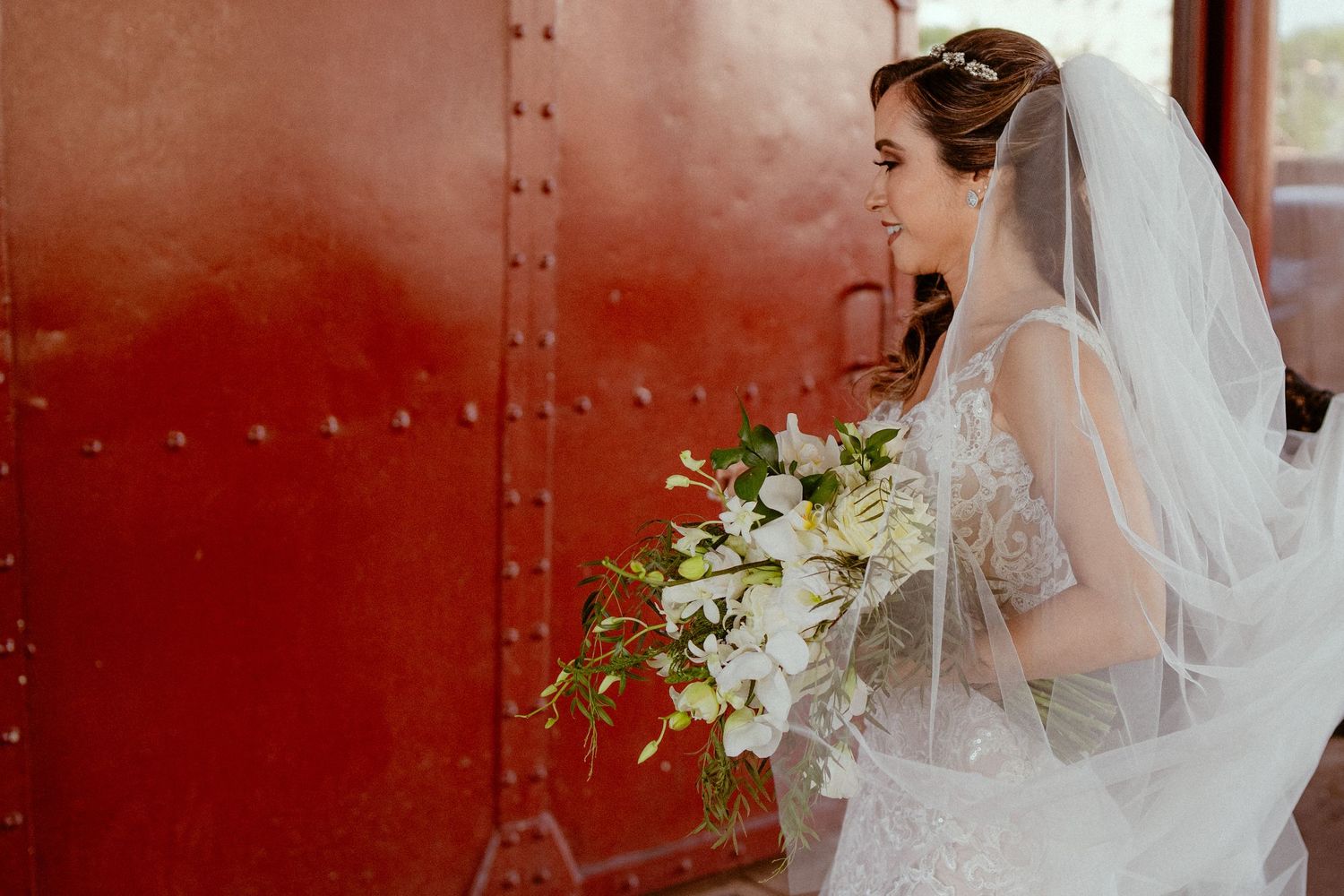Bride stands against a rustic red metal wall while holding a white floral bouquet on her wedding day.