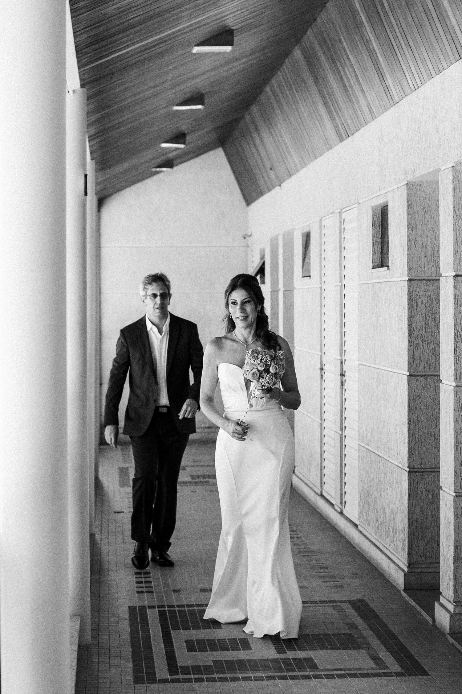 A couple in formal attire walks down an elegant hallway with wood paneled ceiling and modern lighting fixtures.