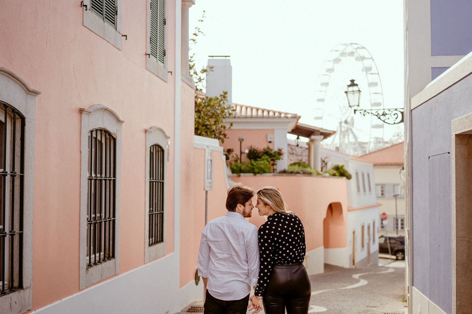 Two people walk down a charming pastel-colored alley in a Mediterranean town with a ferris wheel visible in the distance.