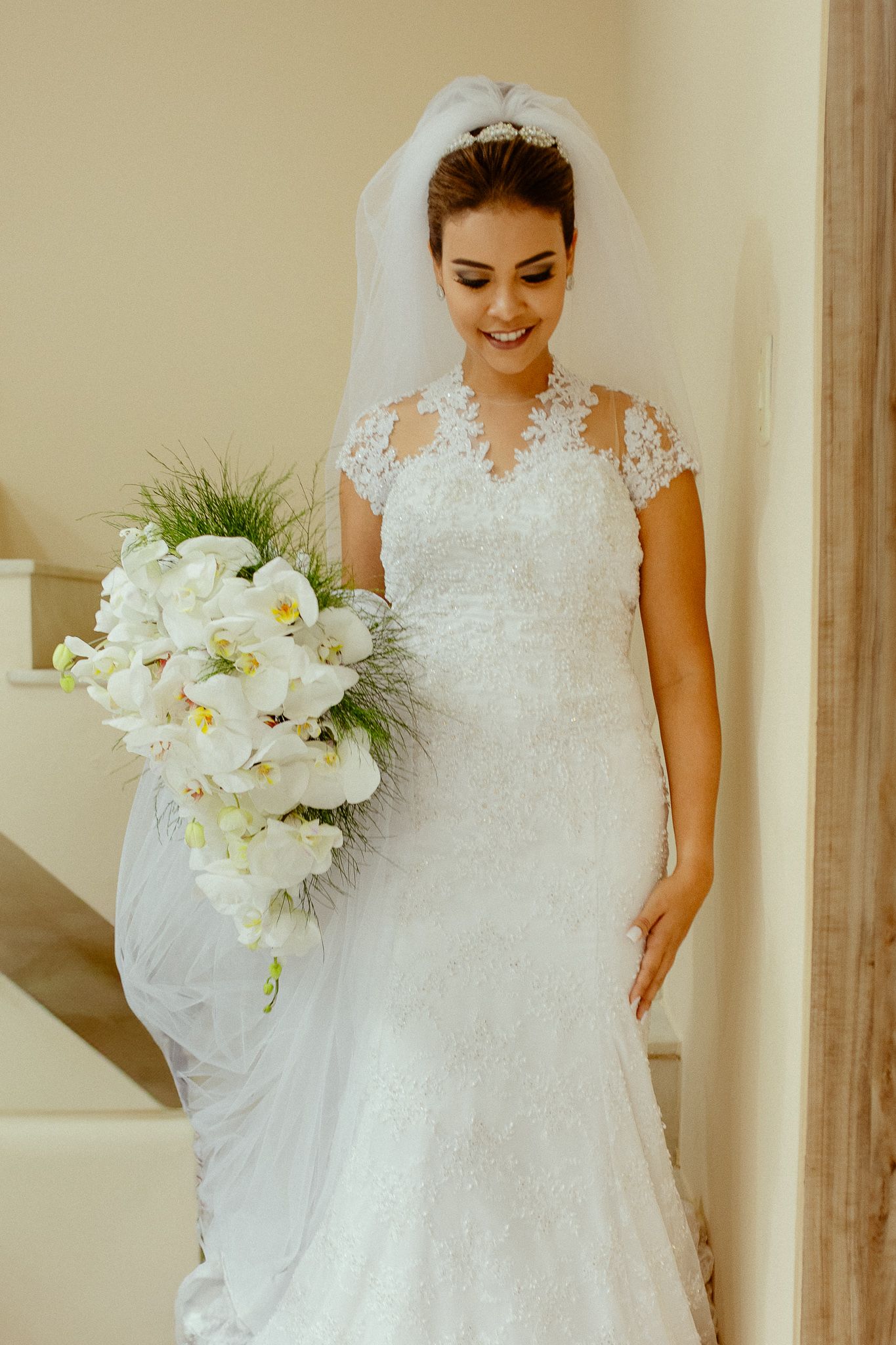 A bride in a white lace wedding dress holds an elegant bouquet of white orchids and flowers against a cream wall.