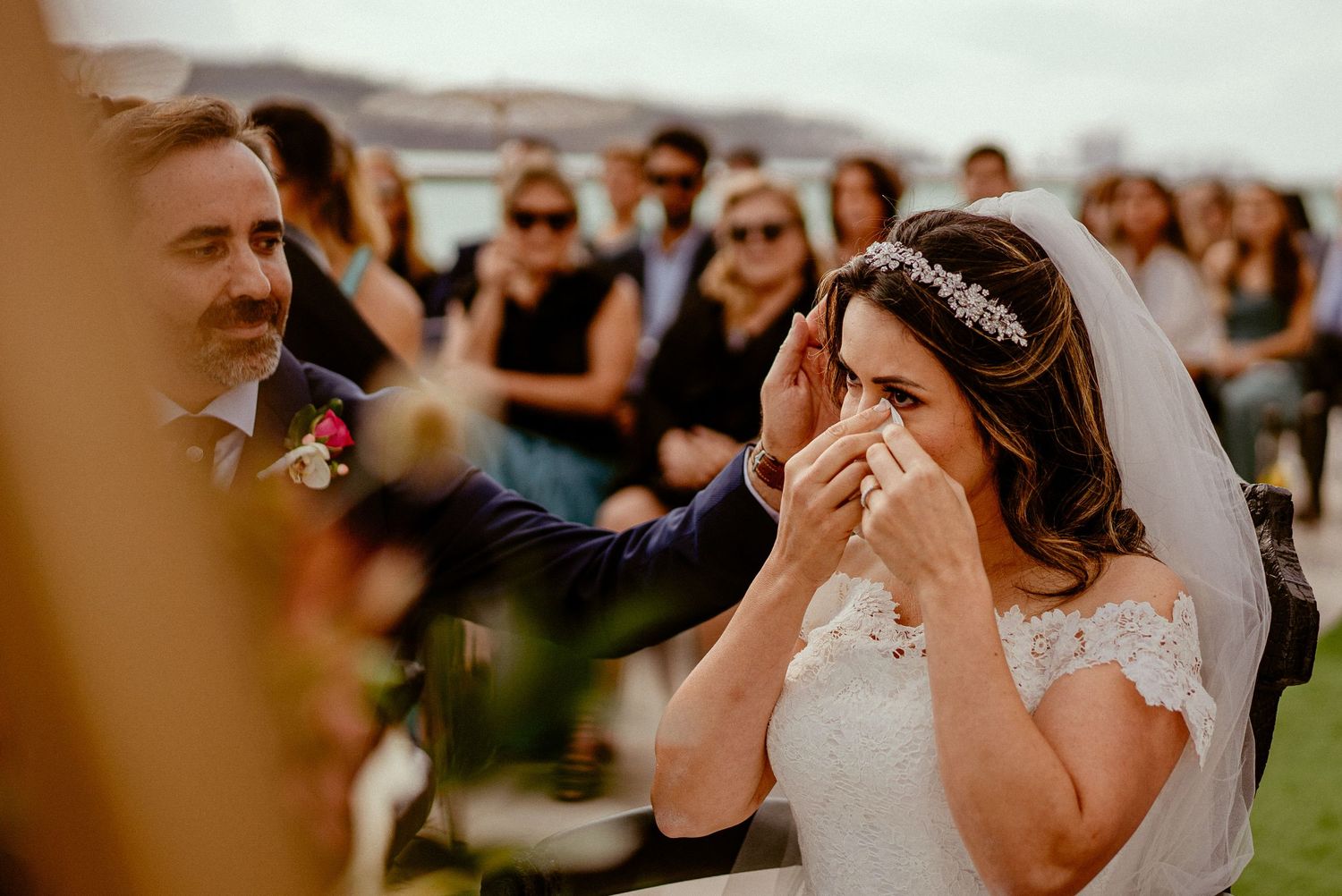 Emotional moment during outdoor wedding ceremony with guests in background.