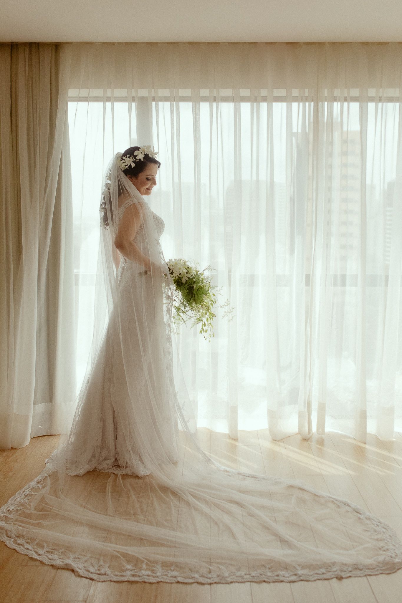 A bride in a flowing white lace wedding gown with cathedral veil stands elegantly by sunlit sheer curtained windows.