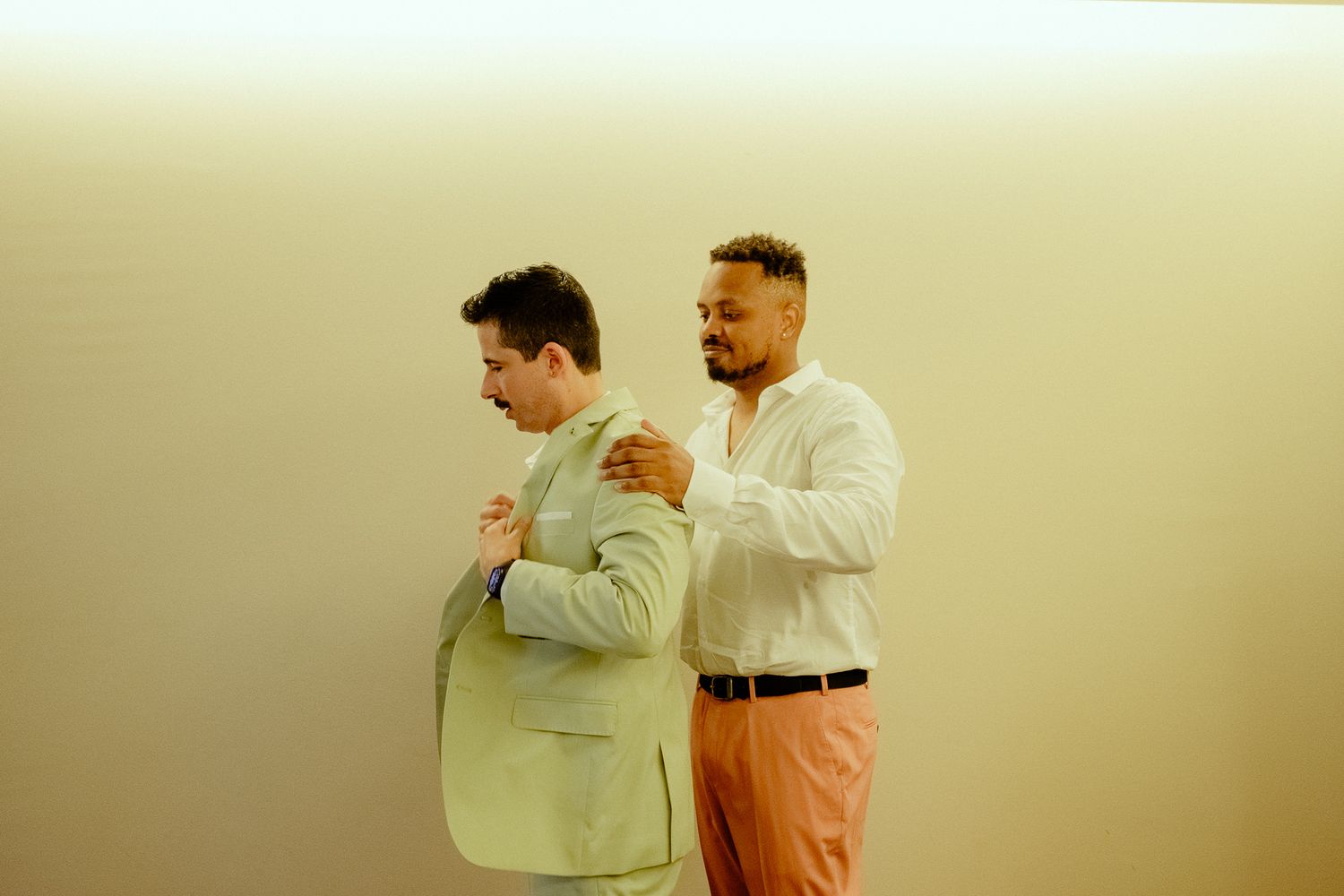 Two men in smart casual attire stand back-to-back, one in a green blazer and one in a white shirt with pink pants.