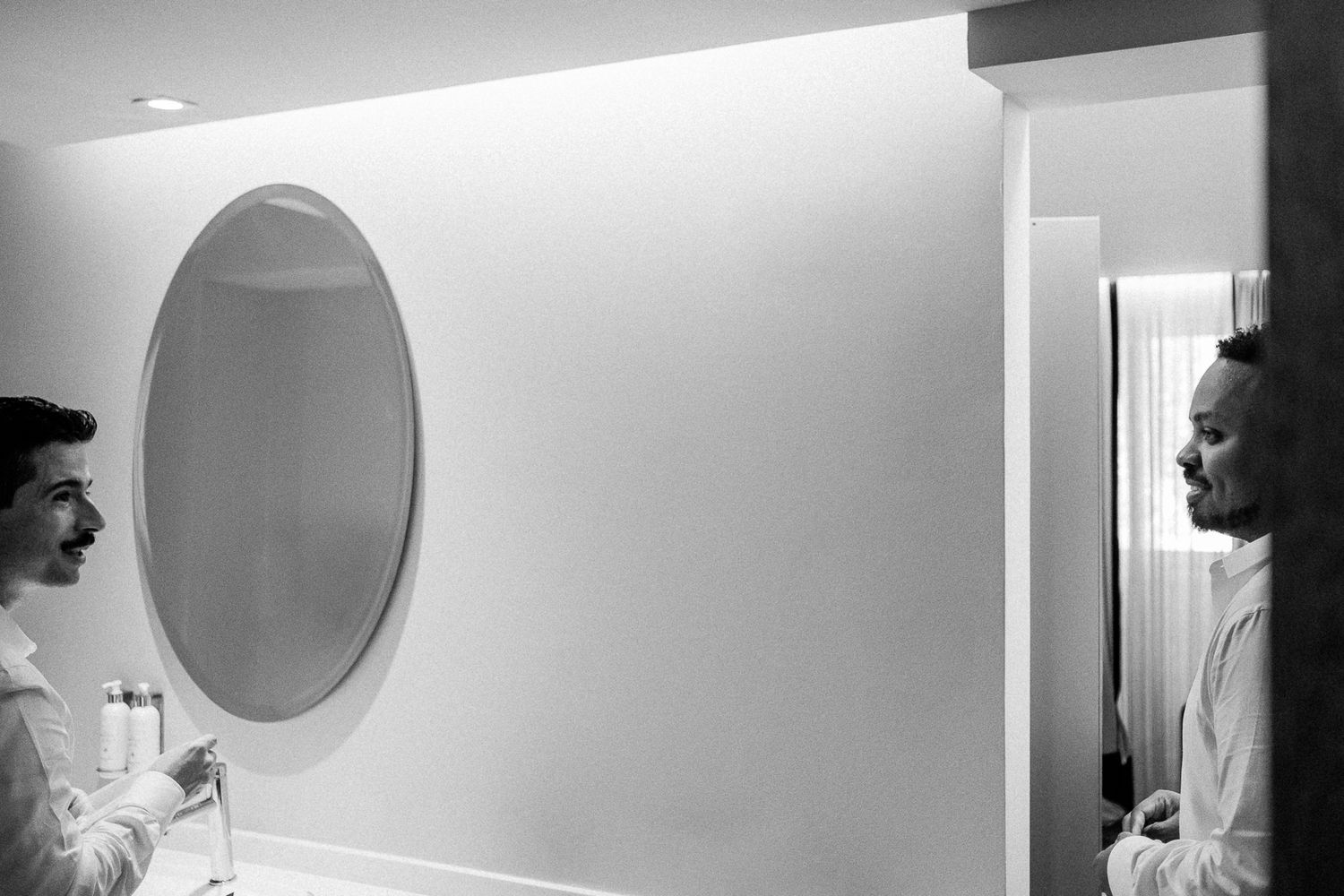 Black and white photo of two men smiling in a bathroom mirror, dressed in white shirts for a wedding.