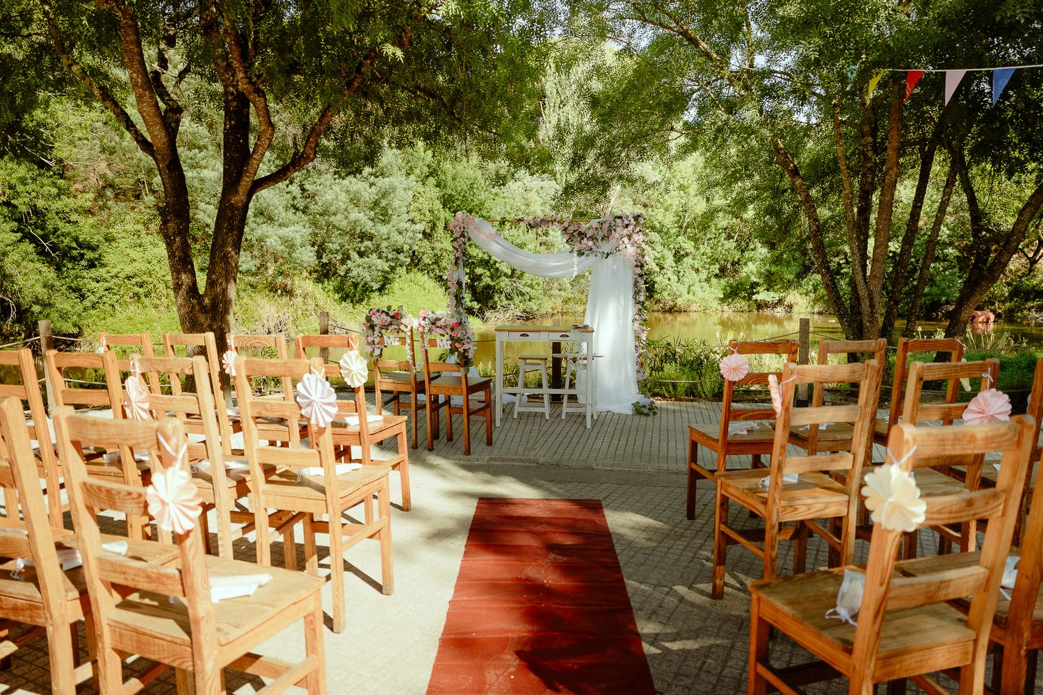 Outdoor wedding ceremony setup with wooden chairs, red carpet aisle, floral arch, and lush green trees in natural garden venue.