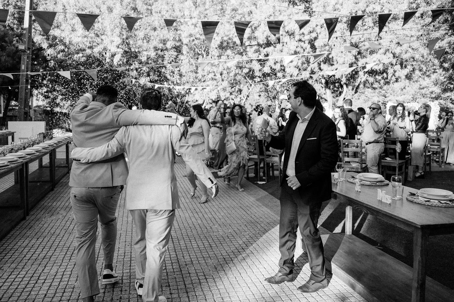Black and white photo of guests celebrating at an outdoor wedding reception with bunting decorations and long dining tables.