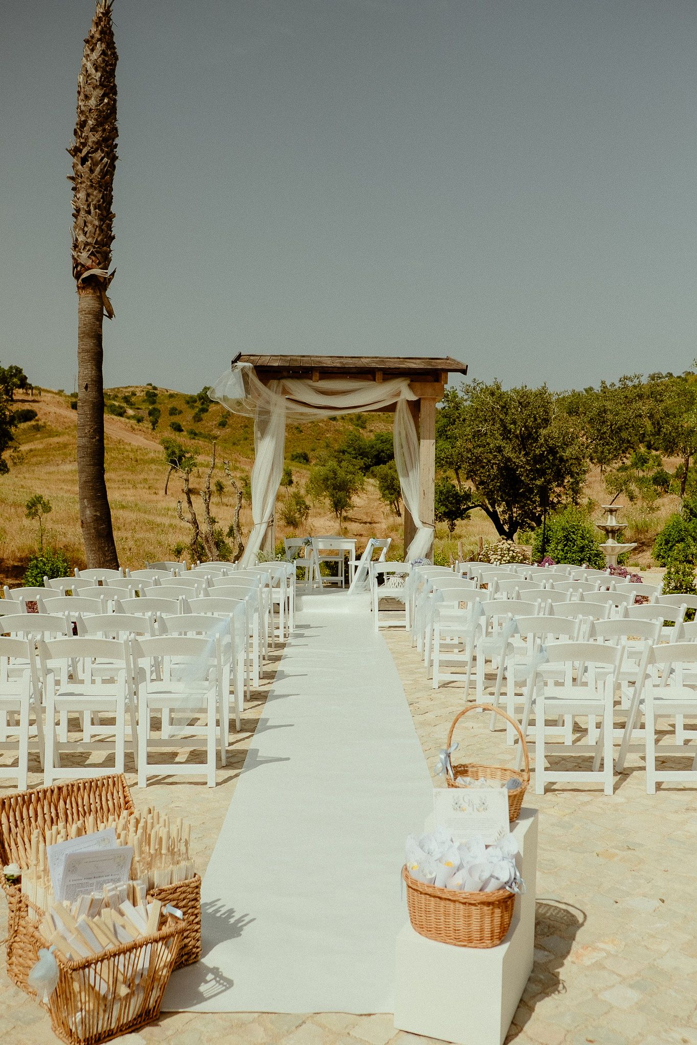 Outdoor wedding ceremony setup with white chairs, aisle runner, wooden arch, and wicker baskets under clear sky.