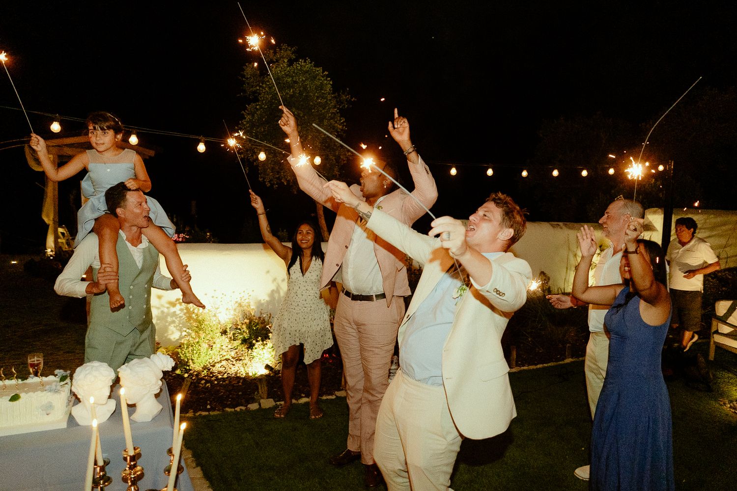 Guests celebrating at an outdoor wedding reception at night, holding sparklers under string lights.