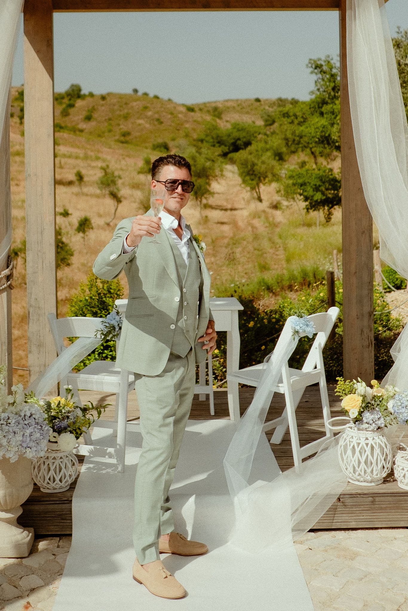 Groom in light green suit holding champagne glass at outdoor wedding ceremony with white chairs and floral decorations.